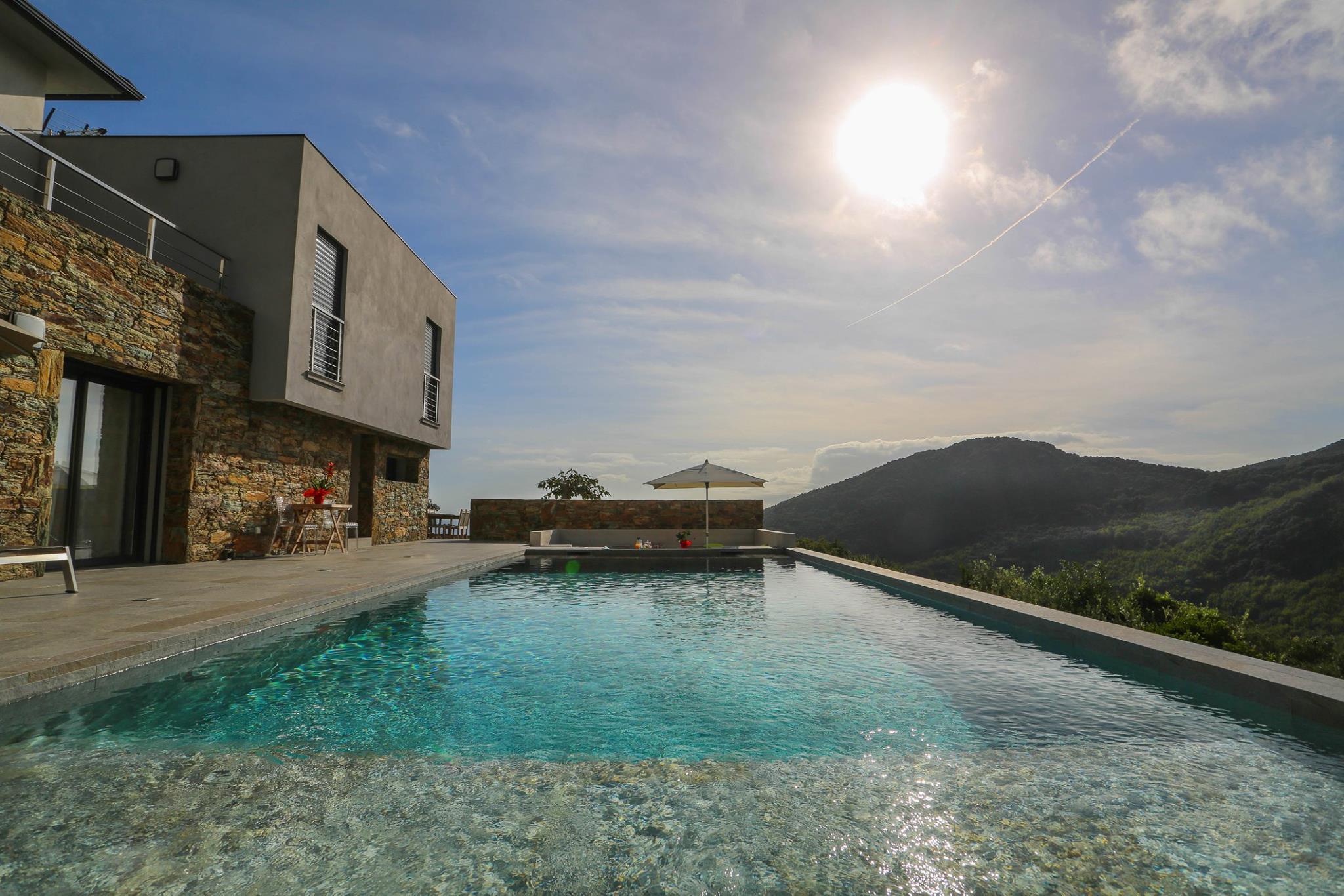 Piscine à débordement turquoise devant une villa en pierre, face au maquis corse