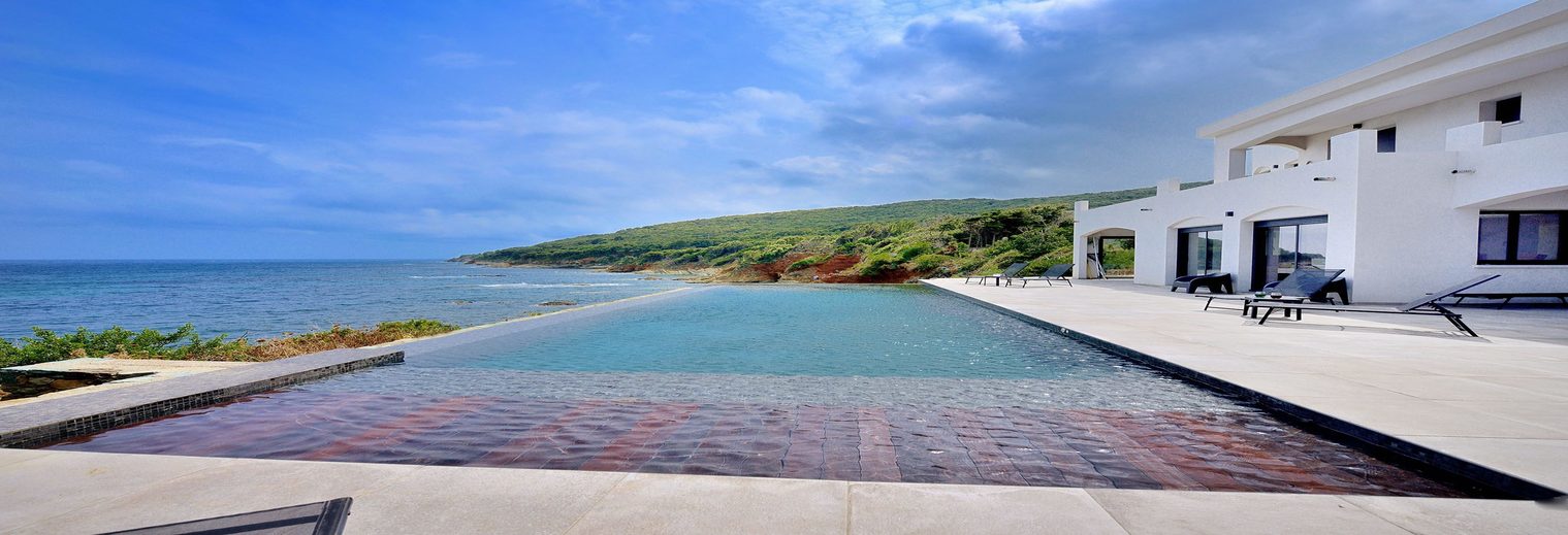 Grande piscine à débordement à fond sombre devant une villa blanche, face à la mer